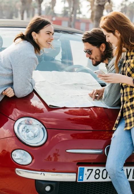 smiling-women-with-smartphone-near-man-looking-map-car-hood
